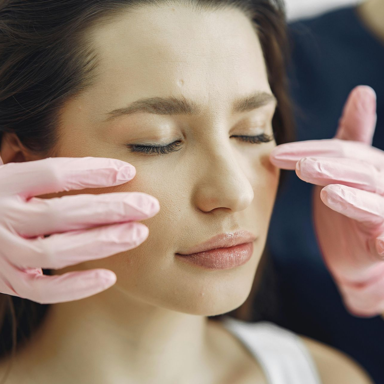 Woman with closed eyes, face being examined by gloved hands, medical setting.